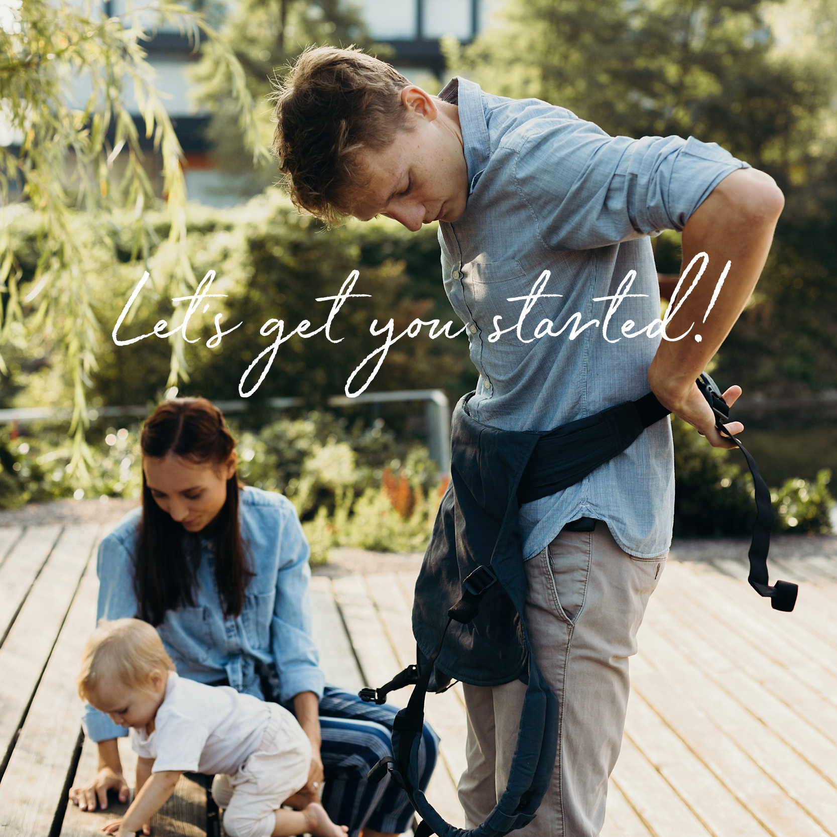 A young blonde dad on a summer trip in nature with his wife and baby girl is standing up and buckling the boba classic carrier around his waist while his daughter is crawling.